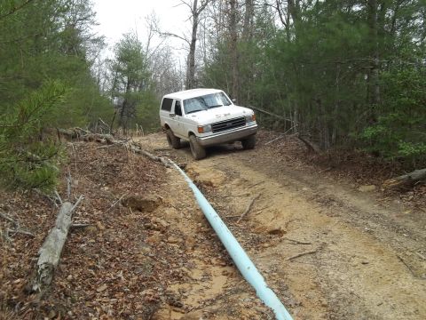 1990 Ford Bronco - A white SUV navigates a rough dirt road through a forest, with a large pipe running alongside the path.