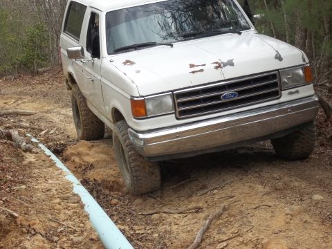 1990 Ford Bronco - A white Ford Bronco navigates a dirt path with a blue pipe alongside.