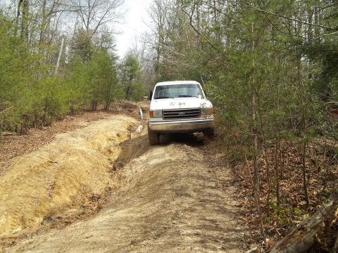 1990 Ford Bronco - A white 1990 Ford Bronco navigates a muddy, rutted dirt path through a wooded area.