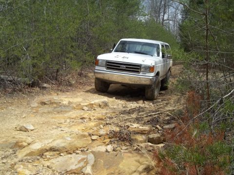 1990 Ford Bronco - A white Ford Bronco navigates a challenging, rocky path through a dense forest.