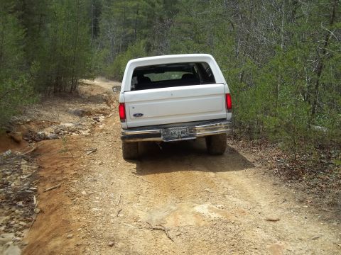 1990 Ford Bronco - White Ford Bronco navigates a rough dirt path through a wooded area.