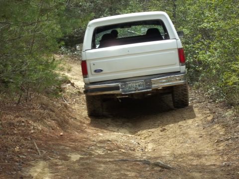 1990 Ford Bronco - A white Ford Bronco navigates a muddy, wooded trail from a rear perspective.