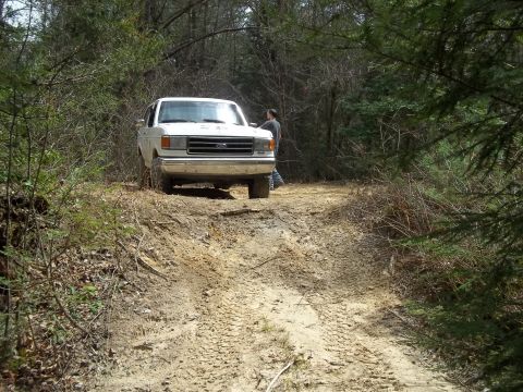 1990 Ford Bronco - A white Ford F-Series truck navigates a muddy, wooded trail.