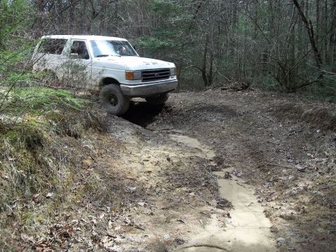 1990 Ford Bronco - White SUV navigates a challenging off-road trail through a wooded area.