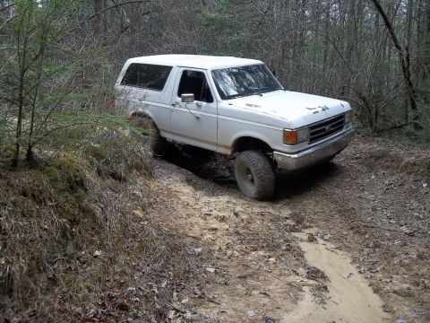 1990 Ford Bronco - White 1990 Ford Bronco navigates a muddy off-road path surrounded by trees.