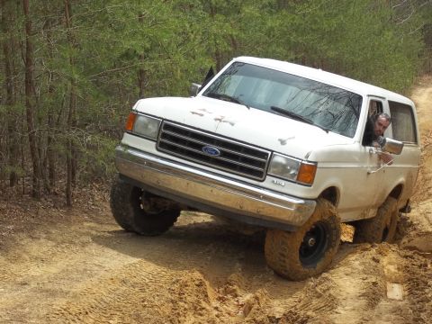 1990 Ford Bronco - A white Ford Bronco navigates a muddy, off-road trail, with its tires kicking up dirt.