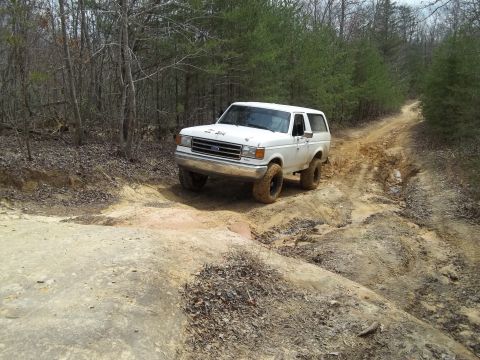 1990 Ford Bronco - A white 1990 Ford Bronco navigates a challenging dirt path through a wooded area.
