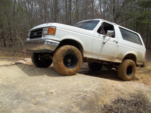 1990 Ford Bronco - White 1990 Ford Bronco navigating an off-road trail with muddy tires.
