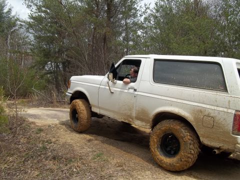 1990 Ford Bronco - A white Ford Bronco navigates a muddy off-road trail with a driver visible inside.