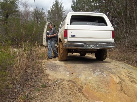 1990 Ford Bronco - White 1990 Ford Bronco is seen off-roading on a rocky, wooded trail with the driver's door open.