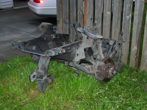 1988 Suzuki Samurai - A disassembled vehicle frame and suspension system, including a brake rotor and caliper, sits on grass next to a wooden fence.