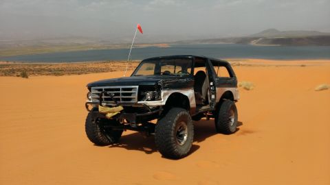 1994 Ford Bronco - This modified Ford Bronco is parked in a desert landscape with sand dunes and a lake in the background.