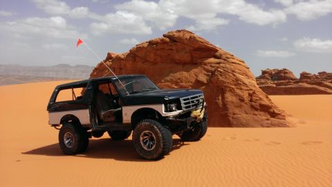 1994 Ford Bronco - This modified Ford Bronco is parked in a desert landscape with sand dunes and rock formations.