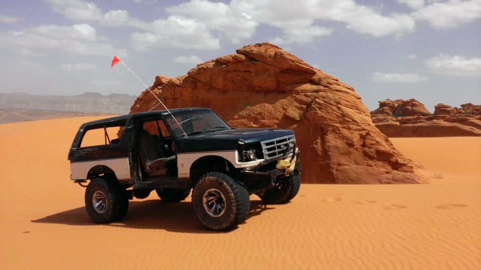 Modified Ford Bronco parked in sand dunes with red rock formations behind it