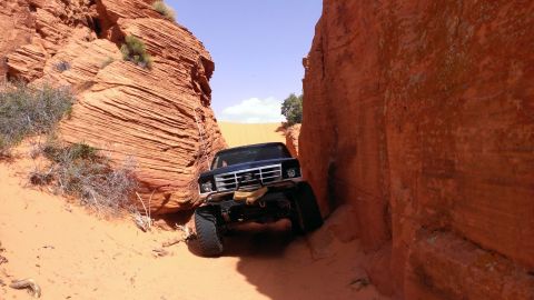 1994 Ford Bronco - Ford Bronco driving through a tight canyon with red rock walls and sandy terrain.