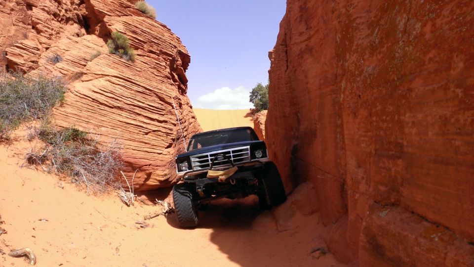 Black Ford Bronco navigating a narrow sandstone canyon with large tires and winch. The vehicle is angled, with its front end visible