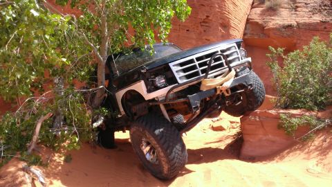 1994 Ford Bronco - This modified truck is shown off-roading on a sandy, rocky trail with a tree on its left.