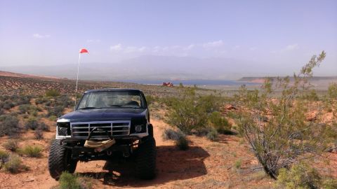 1994 Ford Bronco - A lifted Ford truck with a red flag on a whip antenna sits in a dry, desert environment with a body of water in the background.