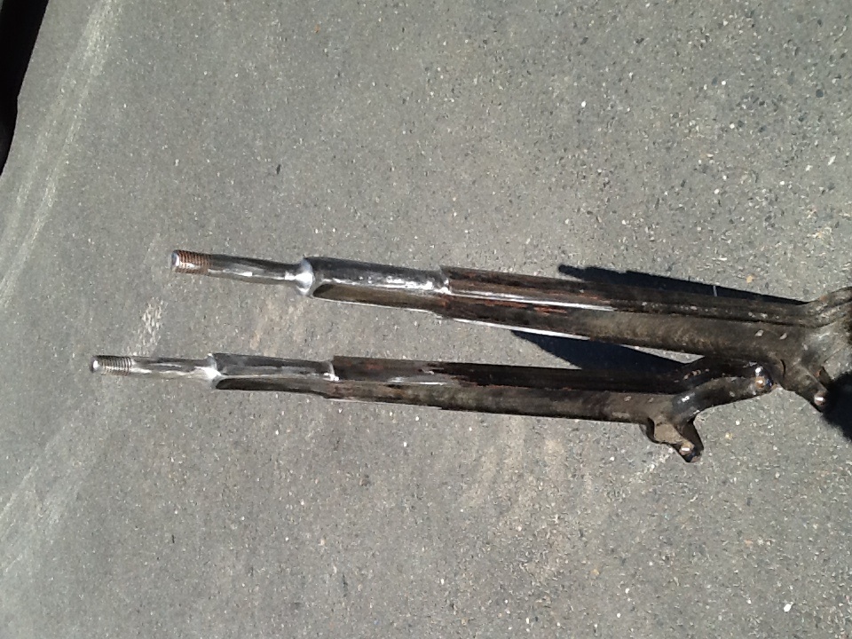 Two bare steel forked radius arms standing upright on asphalt, threaded studs up