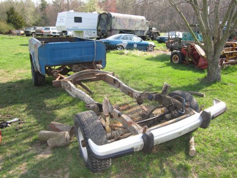 1986 Jeep Grand Wagoneer - Scrapyard scene with a truck chassis in the foreground and other vehicles and an RV in the background.