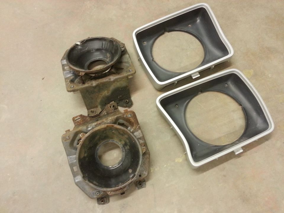 two rusty headlight buckets and two new headlight bezels on a concrete floor