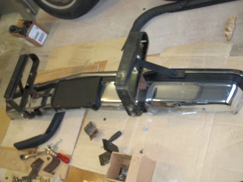 1991 Ford LTD Crown Victoria - A chrome bumper with black trim and associated mounting hardware is laid out on a cardboard-covered floor.