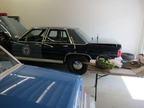 1991 Ford LTD Crown Victoria - A dark-colored police car with its hood open is parked next to a blue car, with a chainsaw on a table nearby.