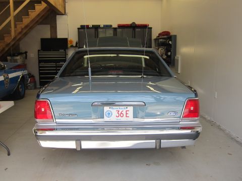 1991 Ford LTD Crown Victoria - The rear of a light blue sedan is shown in a garage, with a police car visible to the left.