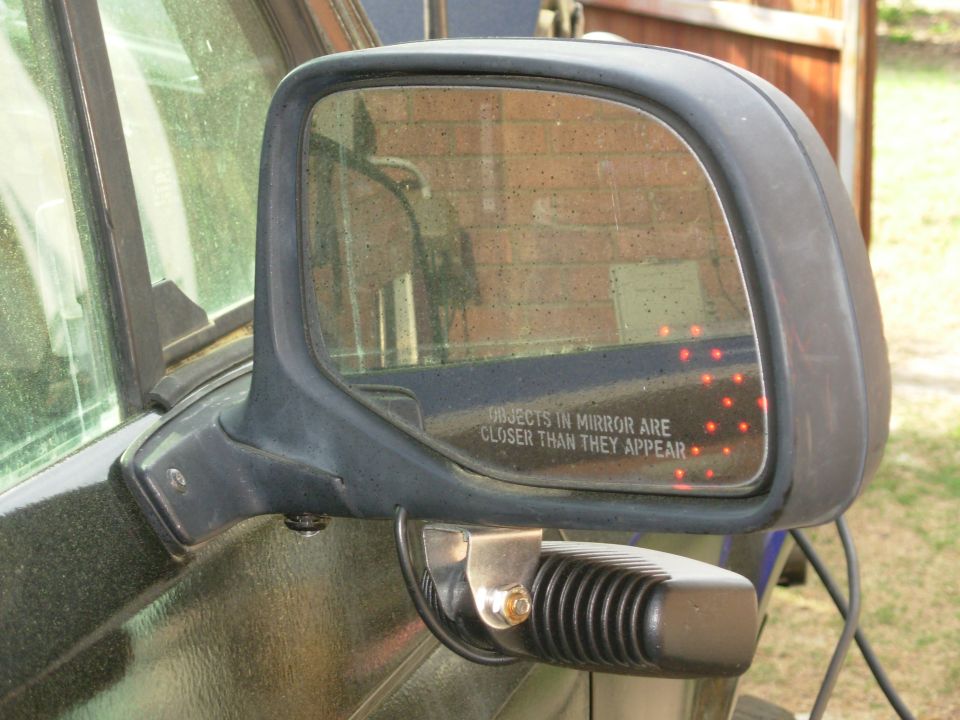 Dark exterior side mirror with lit LED turn-signal arrow and warning text, aftermarket clamp below.