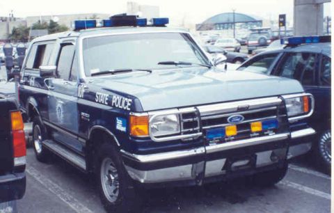 1994 Ford Bronco - A light blue Ford Bronco police SUV is parked in a lot, displaying "STATE POLICE" markings and emergency lights.