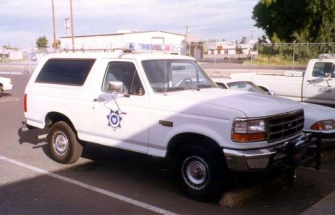 1994 Ford Bronco - A white 1994 Ford Bronco, equipped with police lights, is parked in a lot next to another vehicle.