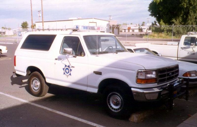 White 1994 Ford Bronco with police lights and a badge on the door, parked next to a pickup truck