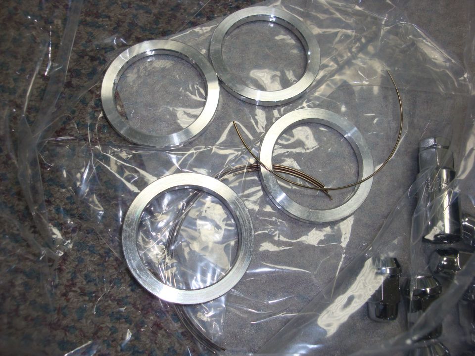 Four aluminum hub-centric rings on plastic wrap with wire clips and chrome lug nuts nearby