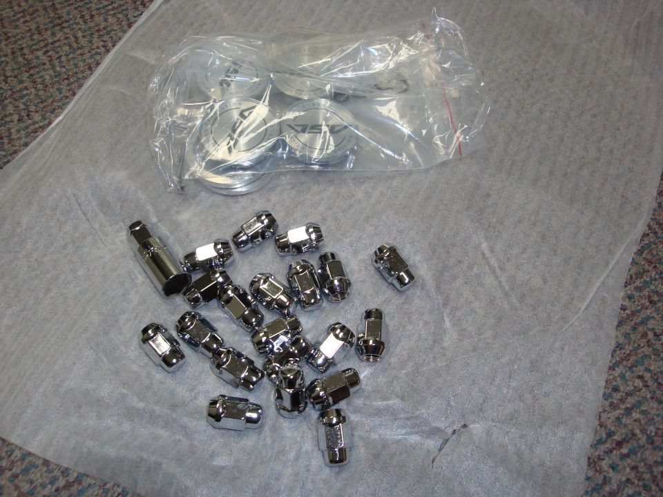 New chrome lug nuts, socket key, and bagged wheel center caps laid out on white tissue paper.