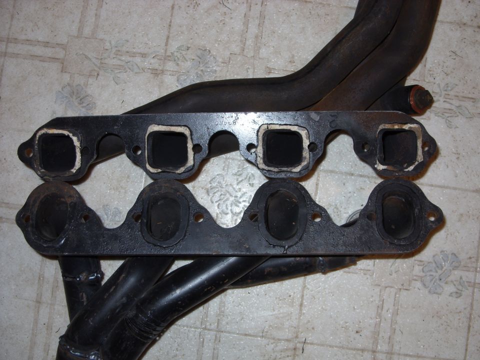 Two black exhaust manifolds with square and oval ports, resting on a tiled floor