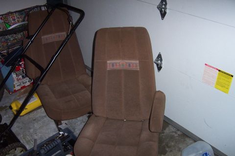 1980 Ford Bronco - A pair of brown cloth high-back bucket seats removed from a vehicle and set on a garage floor against a white wall, each with a woven tree-…