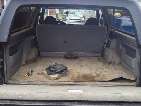 1996 Ford Bronco - The cargo area of the SUV is visible, with a pair of gloves and a toolbox on the floor. Another vehicle is seen through the rear window.