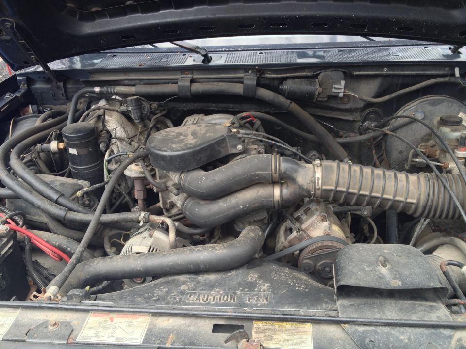 Engine bay with V8 engine, hoses, and fan shroud visible. Text on shroud reads 'CAUTION FAN'