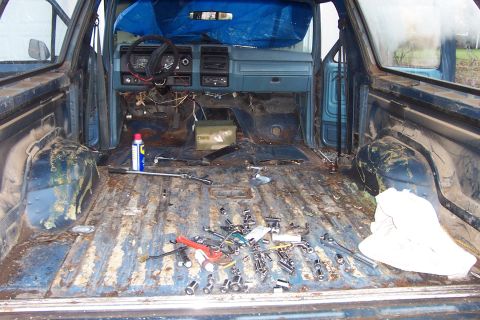1980 Ford Bronco - Interior of a stripped pickup truck cab showing bare rusty floor with scattered tools and hardware, dashboard exposed.