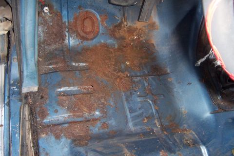 1980 Ford Bronco - Interior floor pan of a blue-painted cab with the carpet stripped out, showing heavy surface rust and flaking debris across the stamped rib…
