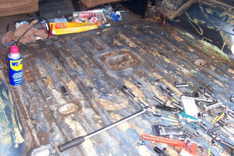 1980 Ford Bronco - Stripped blue rear cargo floor of a truck used as a workbench, with surface rust and old adhesive across the ribbed pan and open body plugs…