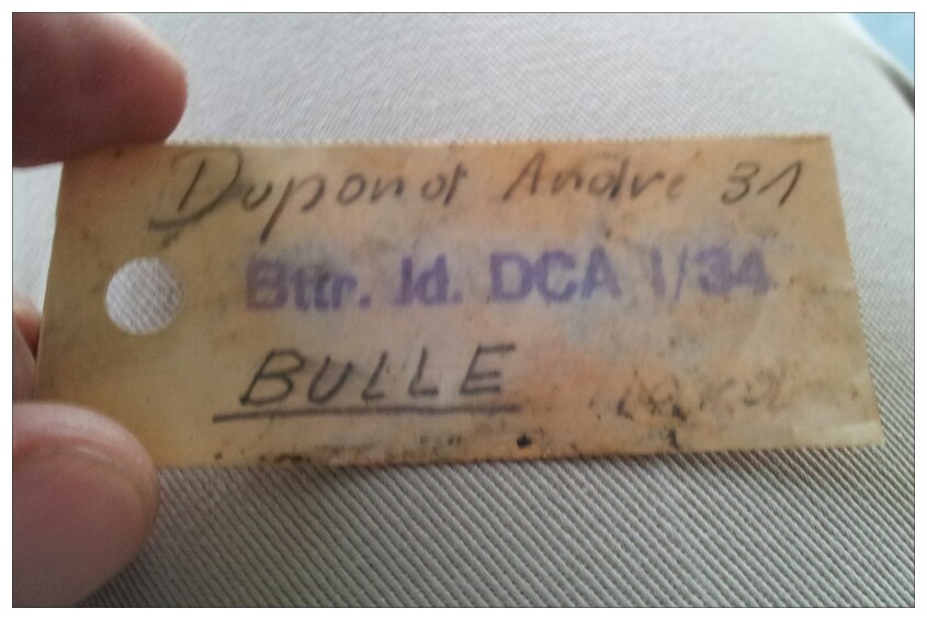 Tan tag with handwritten text: Dupont Andre 31, Btttr. Id. DCA 1/34, BULLE