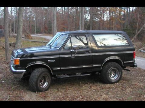 1988 Ford Bronco - Black late-1980s full-size Ford Bronco parked on leaf-covered ground in bare winter woods, three-quarter front view showing the removable h…