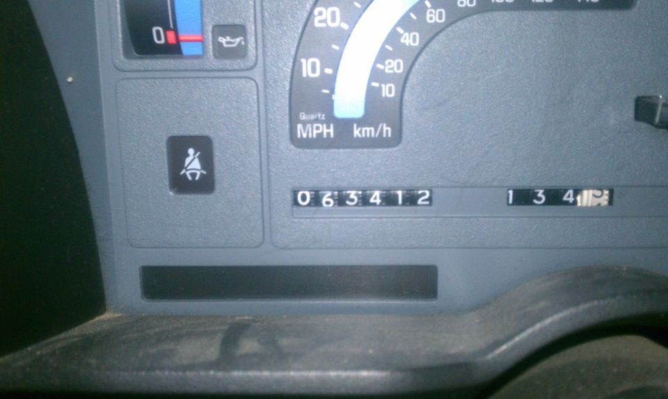 Close-up of vehicle dashboard gauges showing odometer reading 063412 and trip odometer 134.1. Seatbelt warning light is illuminated