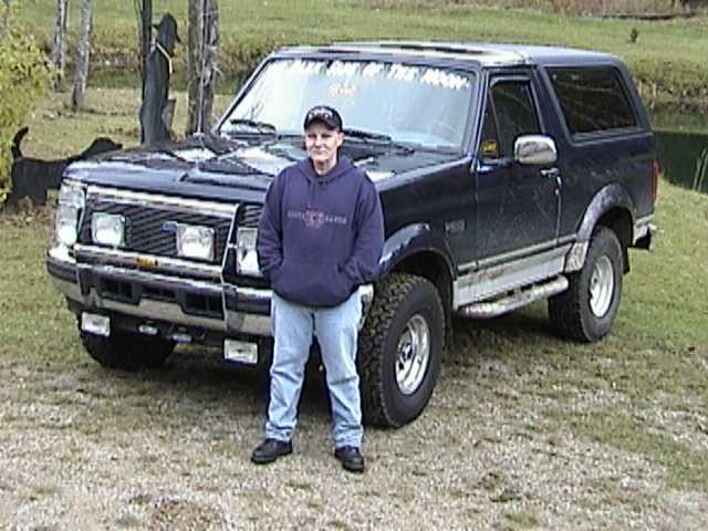 Man standing beside a dark blue lifted Ford Bronco with windshield banner and brush guard