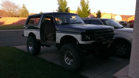 1994 Ford Bronco - This lifted 1994 Ford Bronco, a two-tone paint scheme and open doors, is parked in a driveway next to another vehicle.