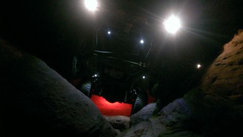 1994 Ford Bronco - An off-road vehicle is seen from a low angle, illuminated by overhead lights and red underglow, as it traverses a rocky landscape at night.