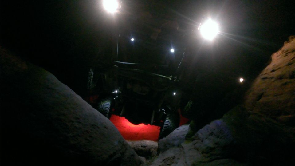 Off-road vehicle with red underglow, illuminated by overhead lights, navigating a rocky, dark terrain