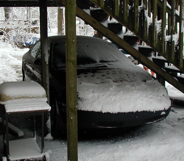 Black sedan parked under wooden stairs, covered in snow. Grill and hood are obscured by snow