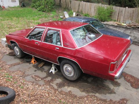 1991 Ford LTD Crown Victoria - A red 1991 Ford Crown Victoria LTD is parked on jack stands in a driveway next to a dark blue car.
